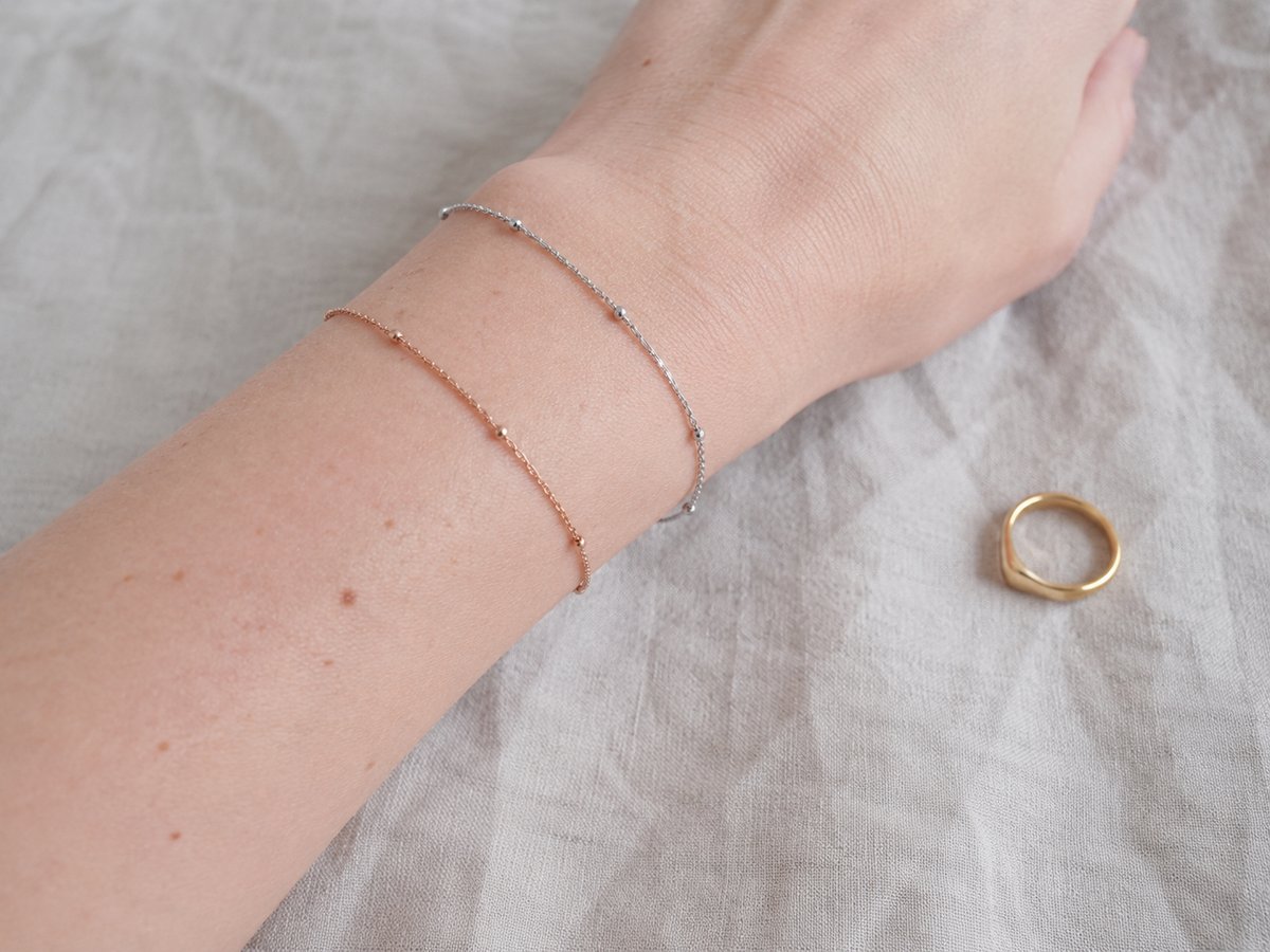 Wrist wearing two delicate bracelets in silver and rose gold, with a small gold ring placed on a beige fabric