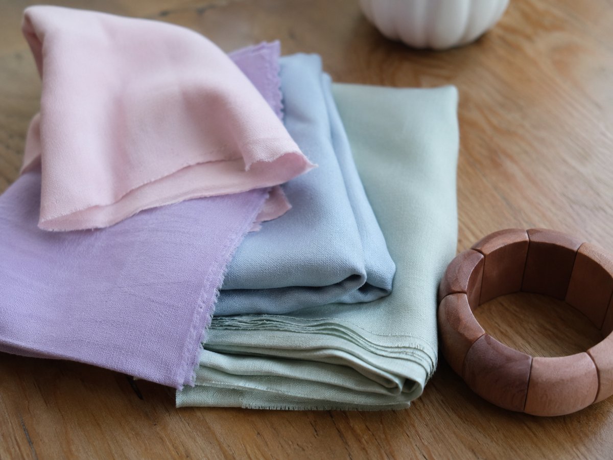 Folded fabric swatches in pastel pink, lilac, powder blue and sage green tones on a wooden surface with a wooden bangle