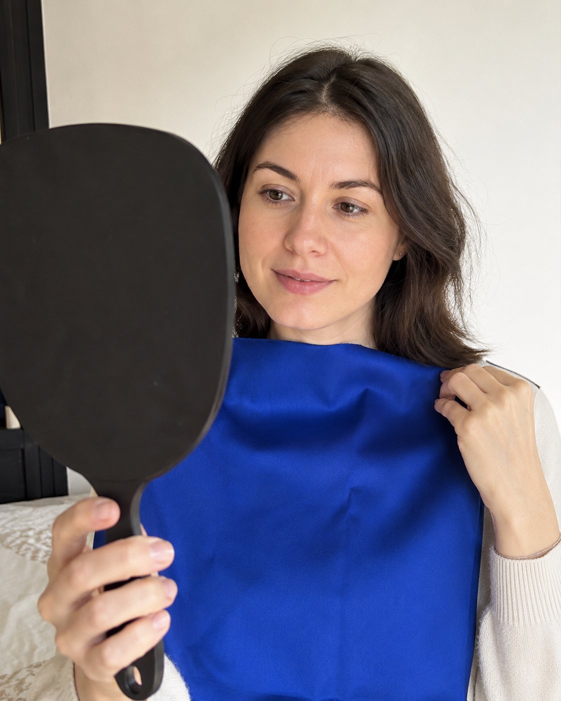 Woman holding a royal blue fabric swatch near her face while looking at a hand mirror, colour draping test
